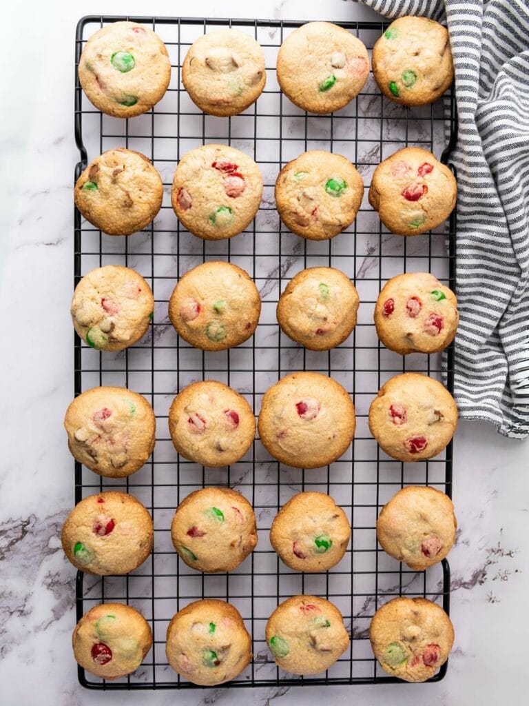 A cooling rack with 28 cookies containing red and green candies, placed on a marble surface next to a striped cloth.