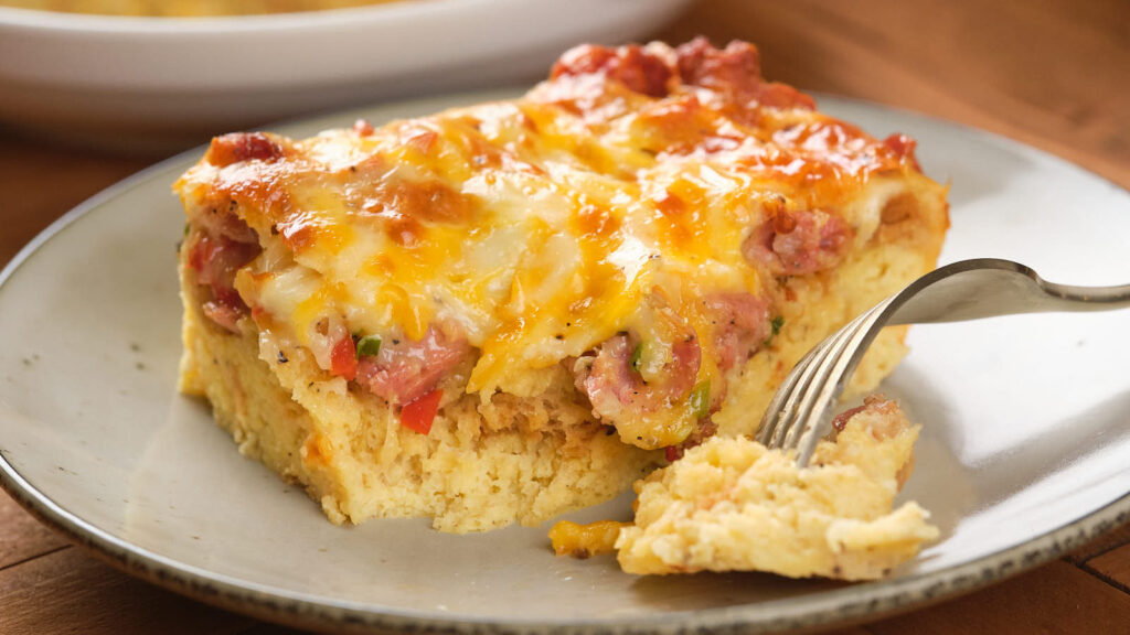 Close-up of a slice of cheesy casserole with bits of meat and vegetables on a plate, with a fork taking a piece.