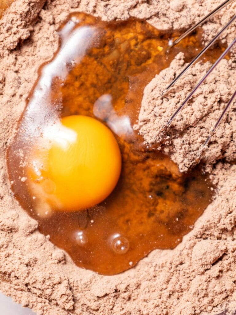 A raw egg yolk on top of brown cake mix, with a whisk nearby.