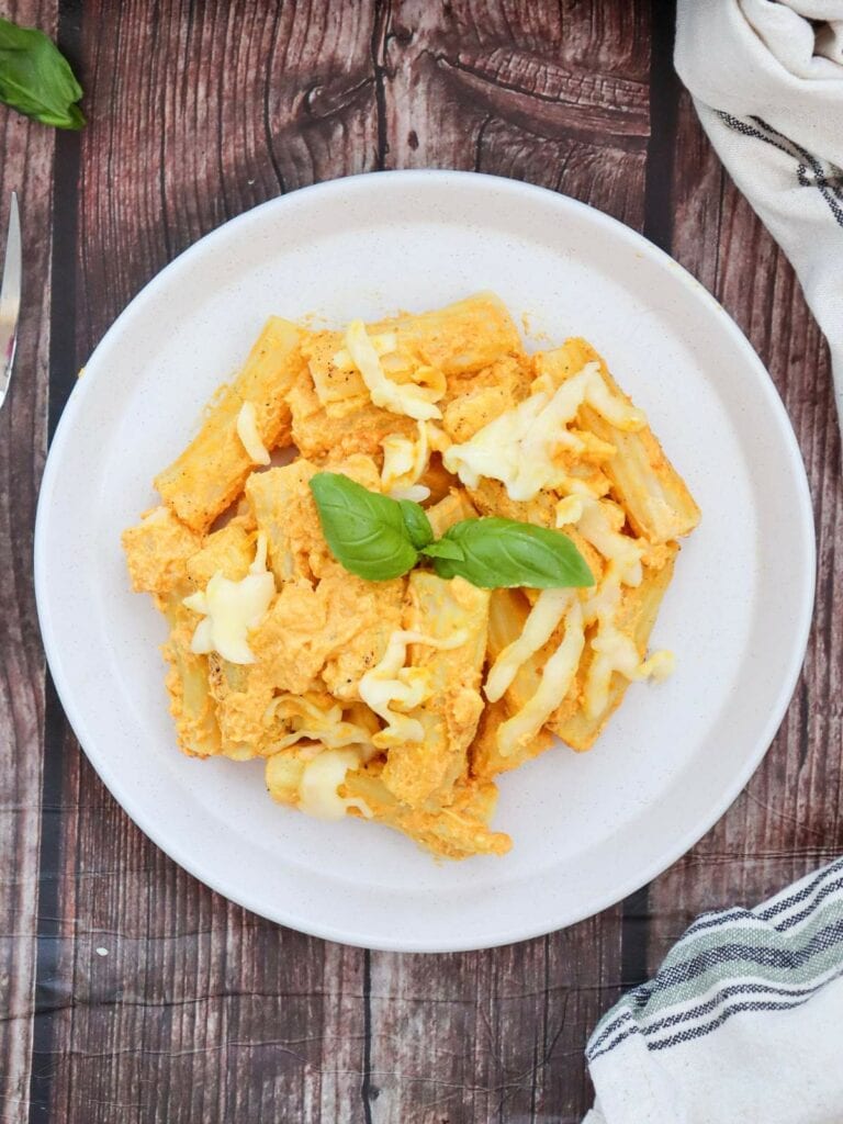 Plate of pumpkin pasta, topped with melted cheese and garnished with fresh basil leaves, on a wooden table.