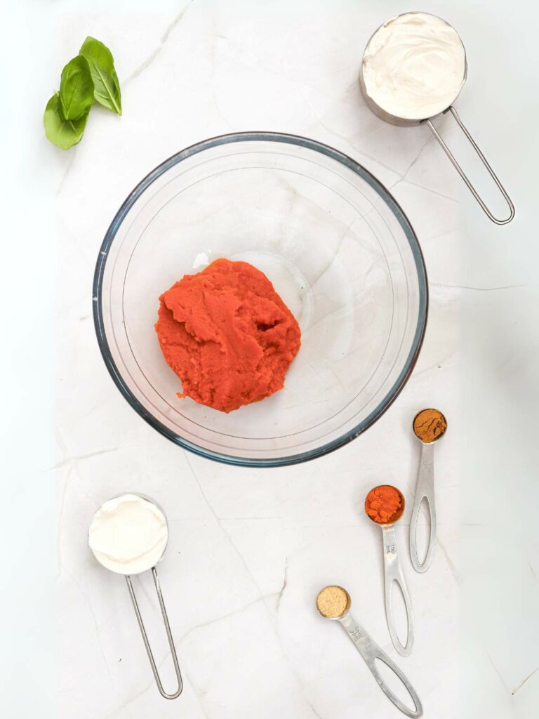 A bowl with red dough is surrounded by measuring cups and spoons holding cream, spices, and herbs on a marble surface.