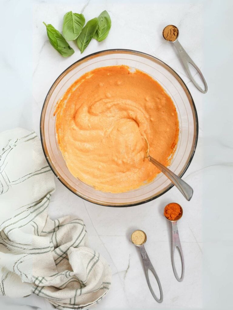 A glass bowl with orange batter, surrounded by basil leaves, measuring spoons with spices, and a cloth on a marble countertop.