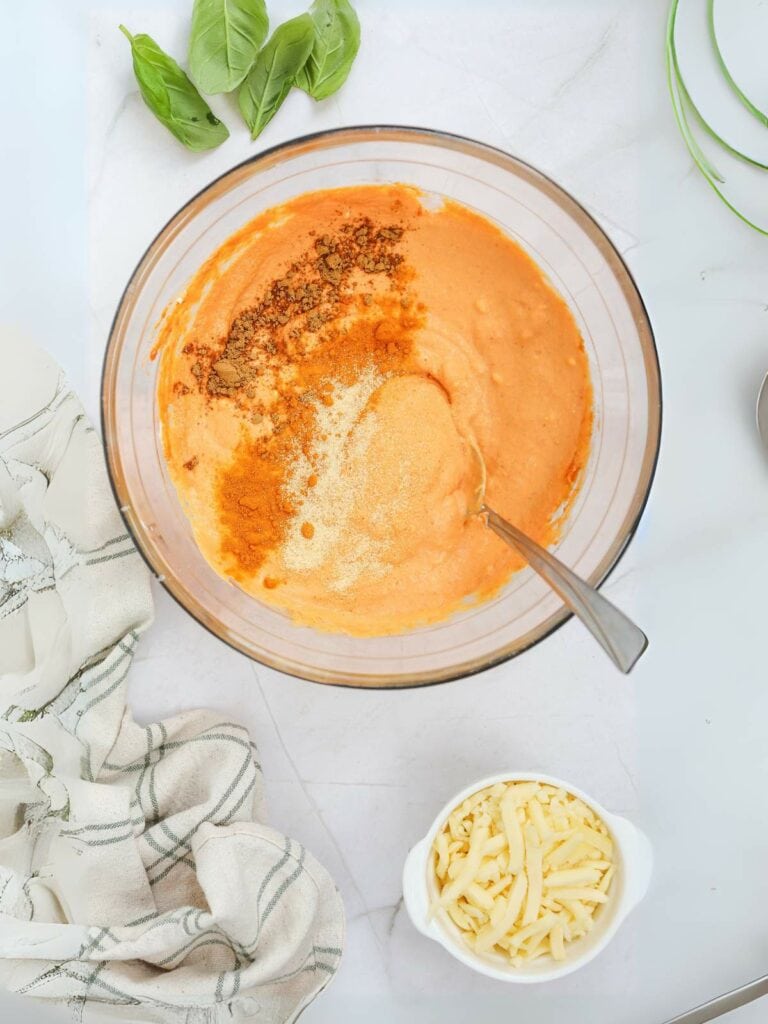 Bowl with orange sauce mixture and spices being stirred, surrounded by fresh basil and a small bowl of shredded cheese on a marble countertop.