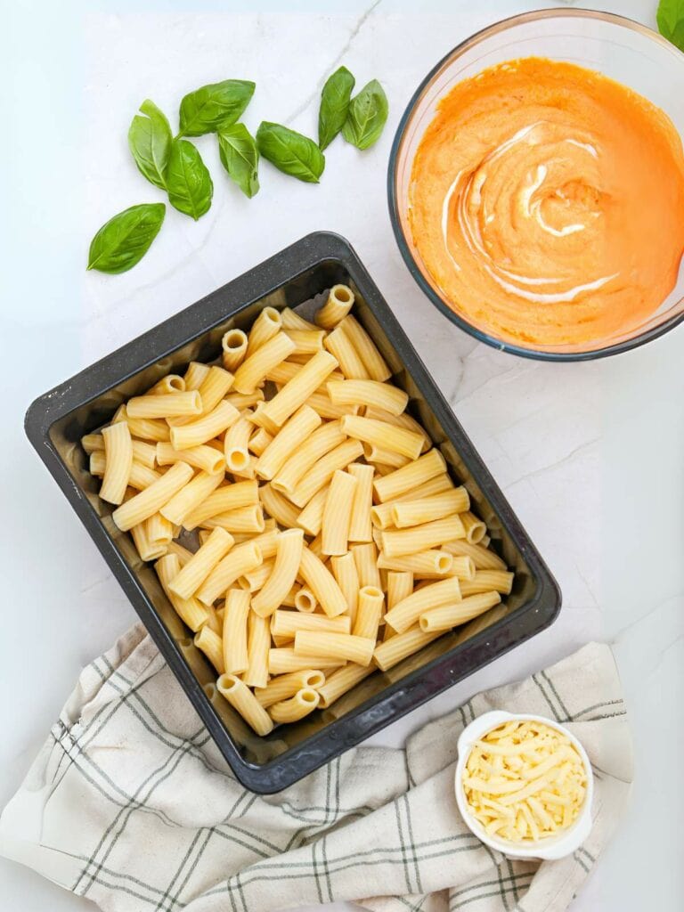 A tray of uncooked rigatoni pasta next to a bowl of creamy orange sauce.