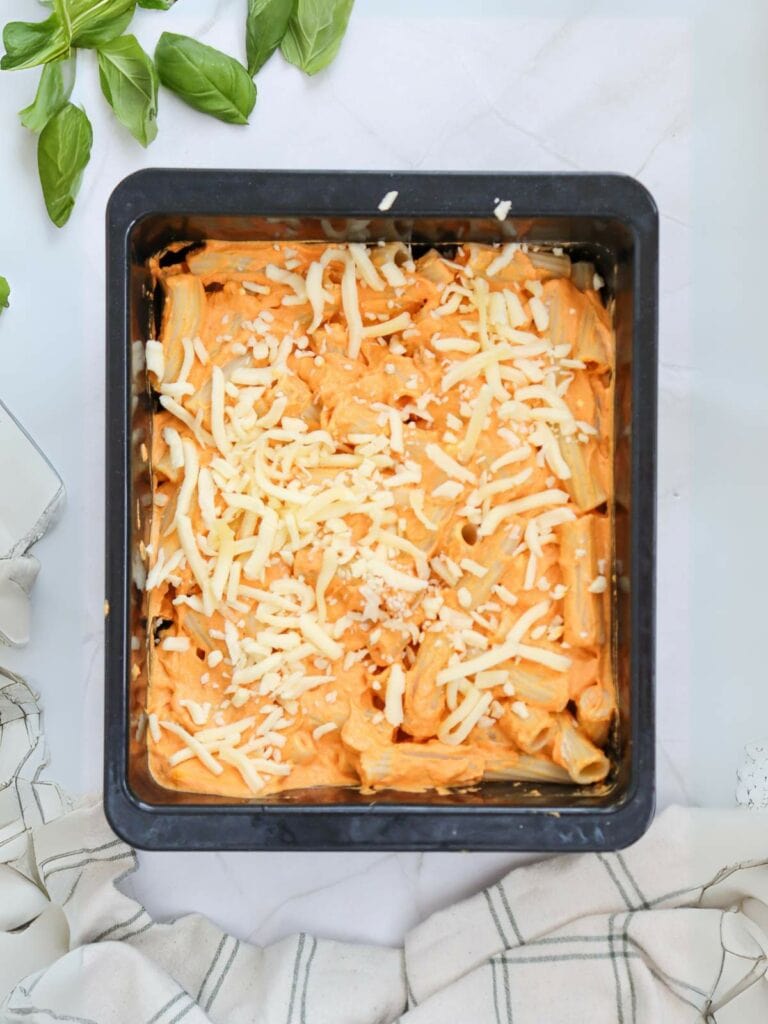 A black square baking dish filled with uncooked cheesy pasta sits on a marble surface.