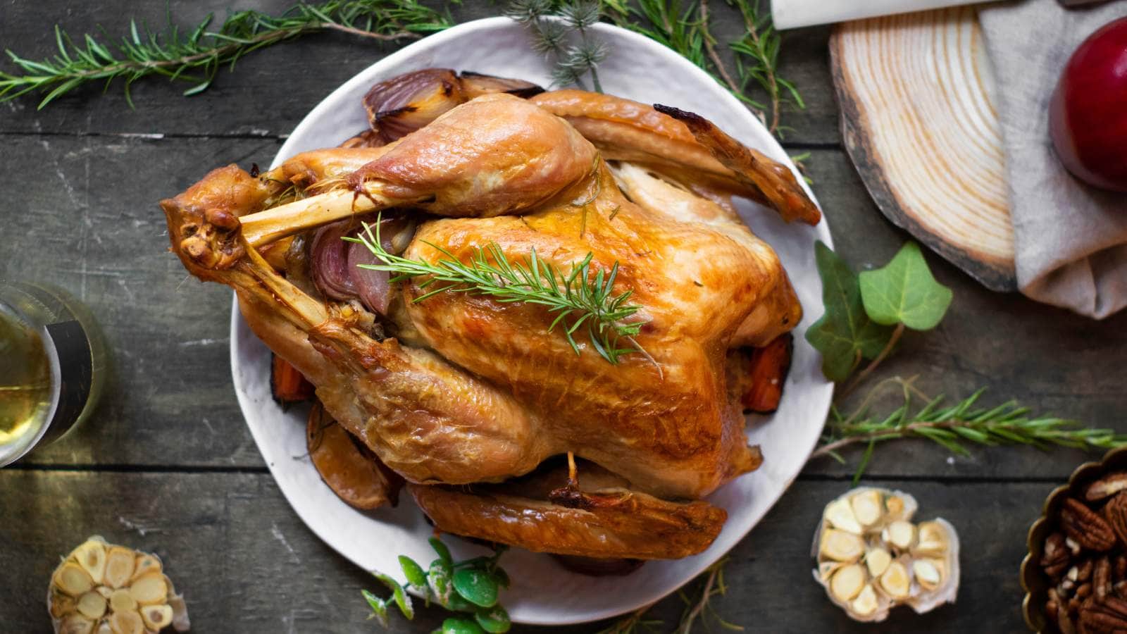 Roasted turkey on a white plate garnished with a sprig of rosemary, surrounded by herbs and sliced garlic on a rustic wooden table.