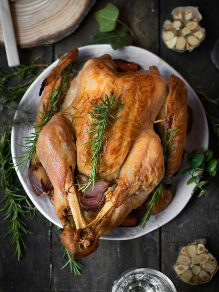 A roasted turkey garnished with rosemary on a white plate, surrounded by garlic and greenery on a dark wooden table.