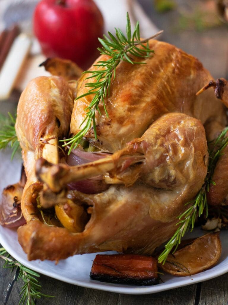 Roasted chicken garnished with rosemary on a white plate, with cooked vegetables and a red apple in the background.