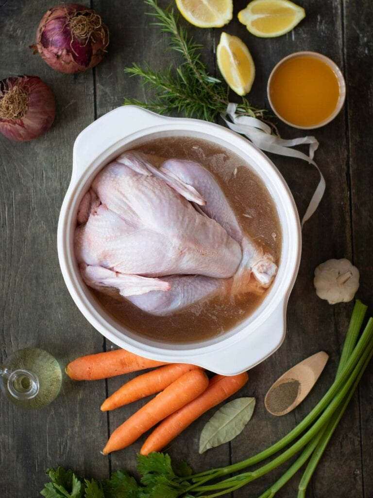 A whole raw chicken in a bowl with broth, surrounded by carrots, onions, lemon, herbs, and a small bowl of liquid.