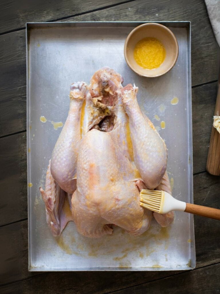 Raw turkey on a baking sheet with a bowl of melted butter and a brush for basting.