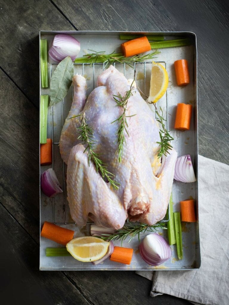 Raw whole chicken on a roasting rack with rosemary, lemon slices, carrot chunks, onion wedges, celery sticks, and bay leaves.