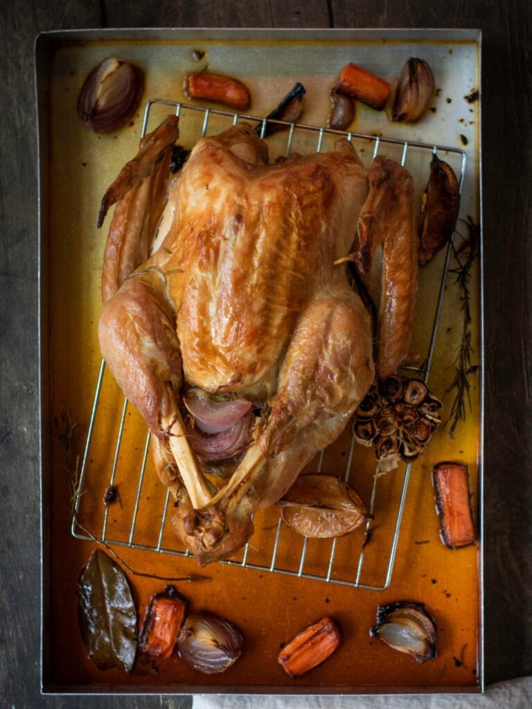 A roasted chicken on a rack in a baking tray, surrounded by roasted vegetables including carrots, onions, garlic, and herbs.