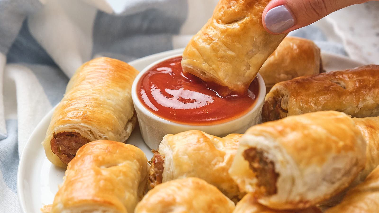 A hand dips a sausage roll into a bowl of ketchup on a plate with several other sausage rolls.