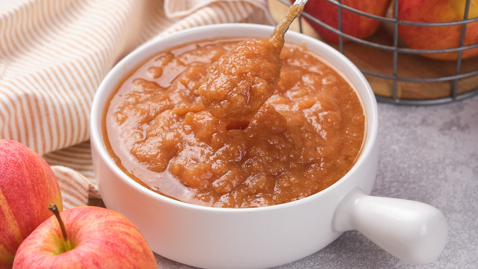 A bowl of applesauce with a spoon, surrounded by apples and a striped cloth on a gray surface.