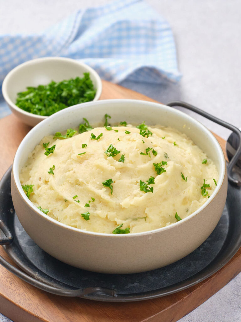 A bowl of mashed potatoes garnished with chopped parsley, placed on a round tray, with a small bowl of additional parsley in the background. A blue checkered cloth is visible nearby.