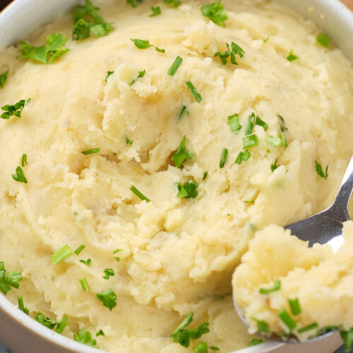 A bowl of creamy mashed potatoes garnished with chopped parsley, with a spoon resting inside.