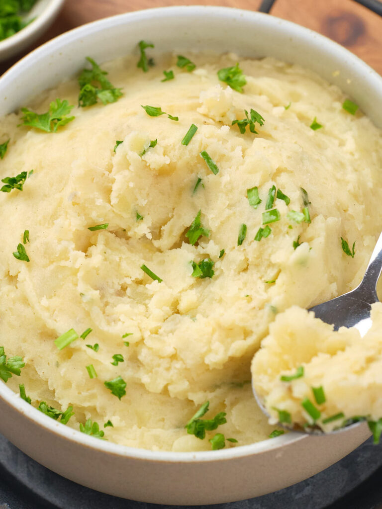 A bowl of creamy mashed potatoes garnished with chopped parsley, with a spoon resting inside.