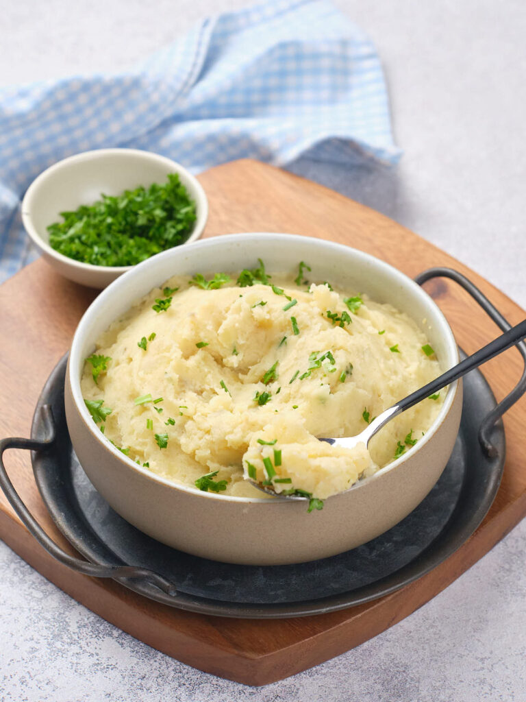 A bowl of mashed potatoes garnished with chopped herbs, placed on a wooden board with a side dish of additional herbs.