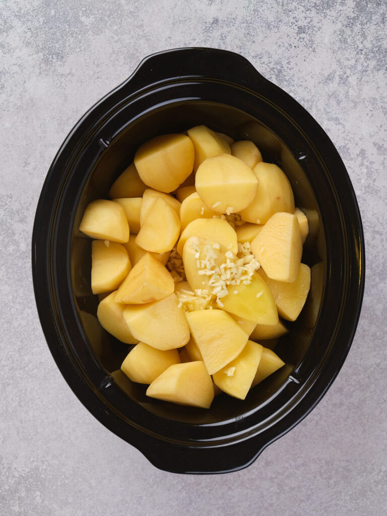 Chopped potatoes and minced garlic in a black slow cooker, placed on a gray surface.