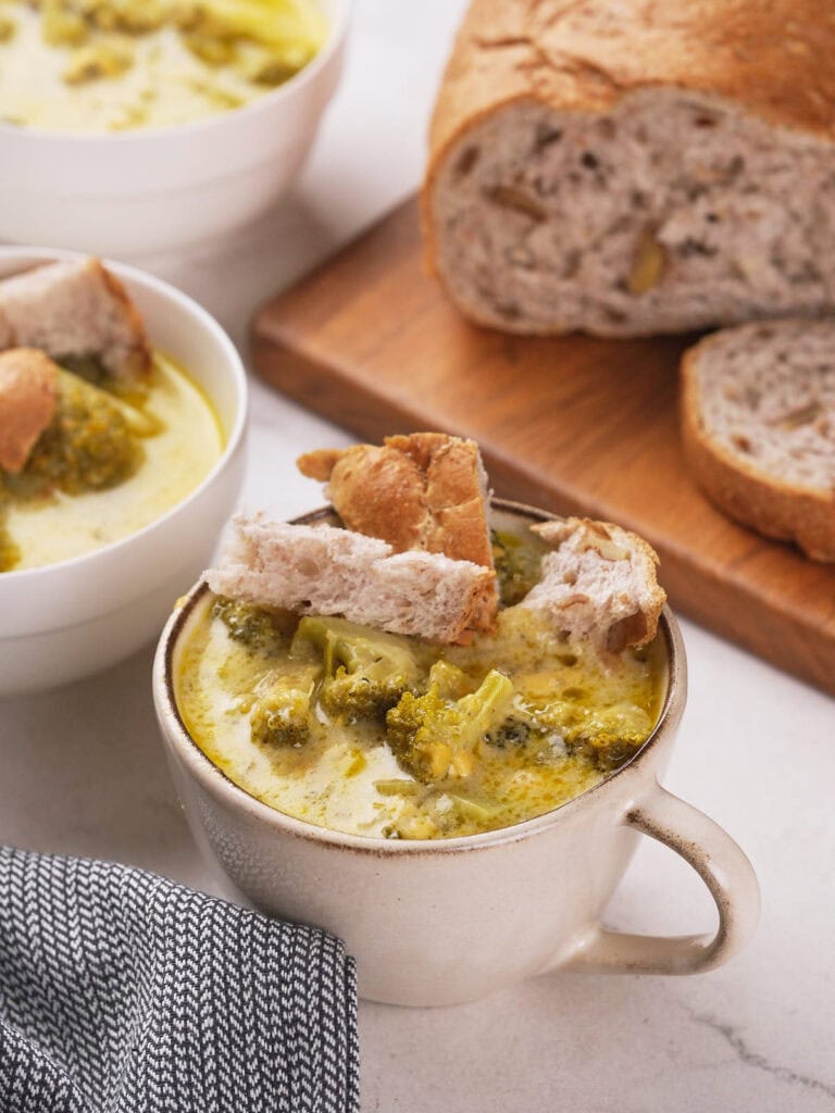 A mug of broccoli cheddar soup topped with bread pieces, accompanied by slices of bread on a wooden board. A bowl of soup is in the background next to a gray cloth napkin.