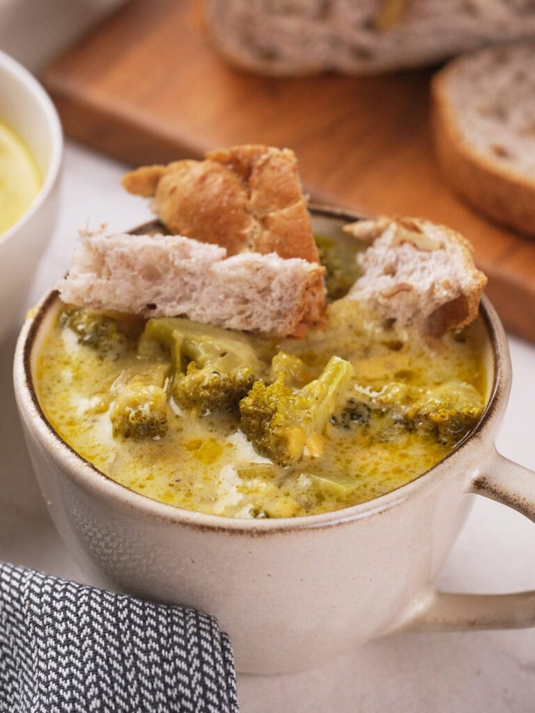 Creamy broccoli cheddar soup in a white mug topped with bread pieces, with a slice of bread on a wooden board.