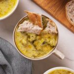 A bowl of broccoli cheddar soup with a piece of bread on top, next to a wooden board with more bread.