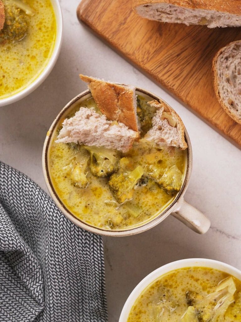 A bowl of broccoli cheddar soup with a piece of bread on top, next to a wooden board with more bread.