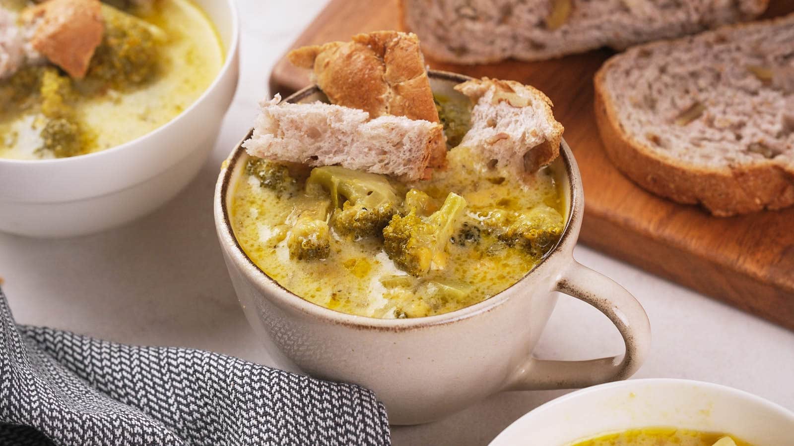 A bowl of broccoli cheddar soup topped with bread pieces next to sliced bread on a wooden board.