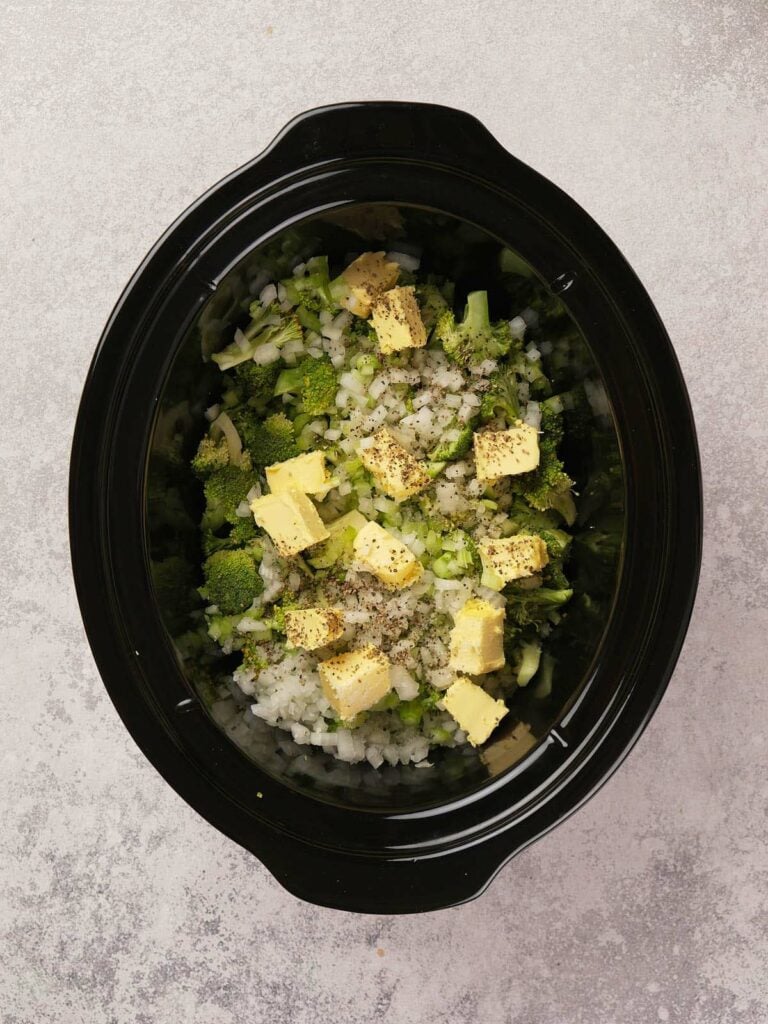 Chopped broccoli, onions, and cubes of butter with pepper in a black slow cooker.