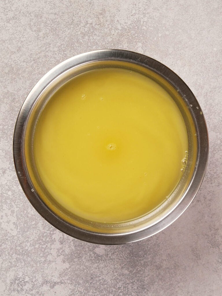 A stainless steel bowl filled with yellow clarified butter on a textured gray surface.