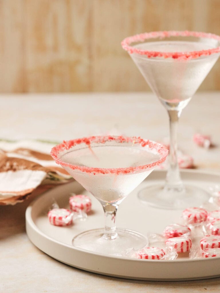 Two candy cane martini glasses with red sugar rims on a tray surrounded by peppermint candies.