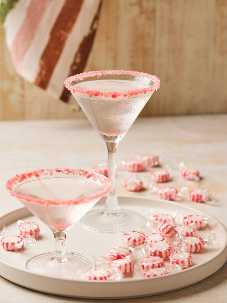 Two candy cane martini on a tray surrounded by wrapped peppermint candies.