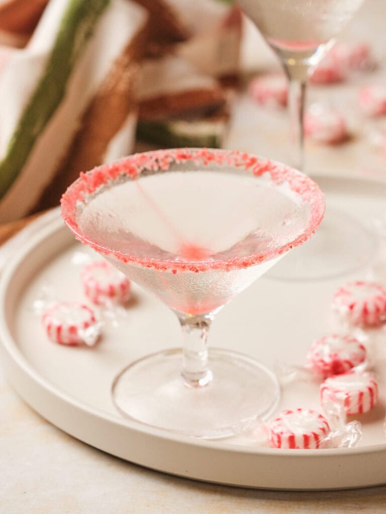 Candy cane martini with a red sugar rim sits on a tray surrounded by wrapped peppermint candies.