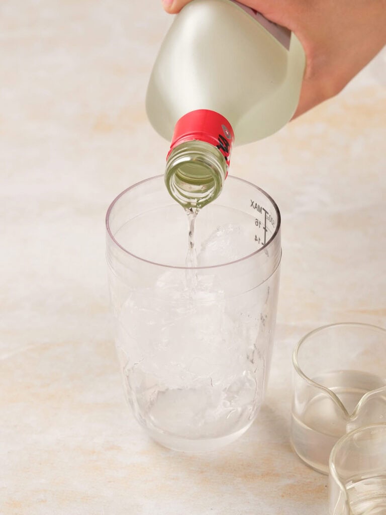 A hand pours clear liquid from a bottle into a transparent container filled with ice.