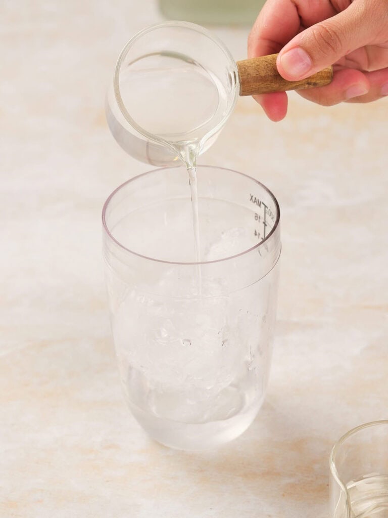 A hand pouring liquid from a measuring cup into a glass filled with ice.