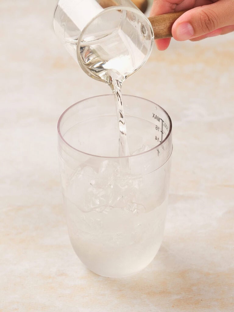 A hand pours liquid from a measuring cup into a clear glass half-filled with ice, on a light-colored surface.