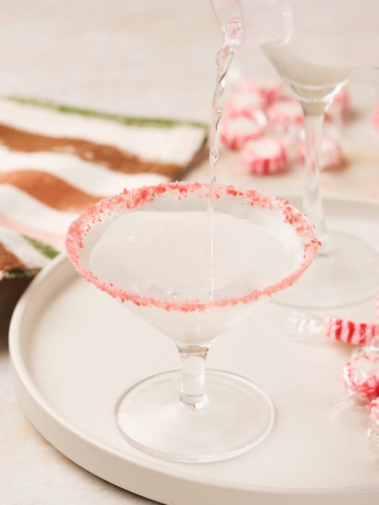 A drink is poured into a sugar-rimmed martini glass on a white tray with peppermint candies nearby.