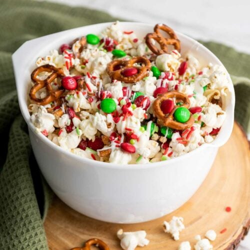 A bowl of popcorn mixed with pretzels, red and green candies, sprinkles, and drizzled with white chocolate.