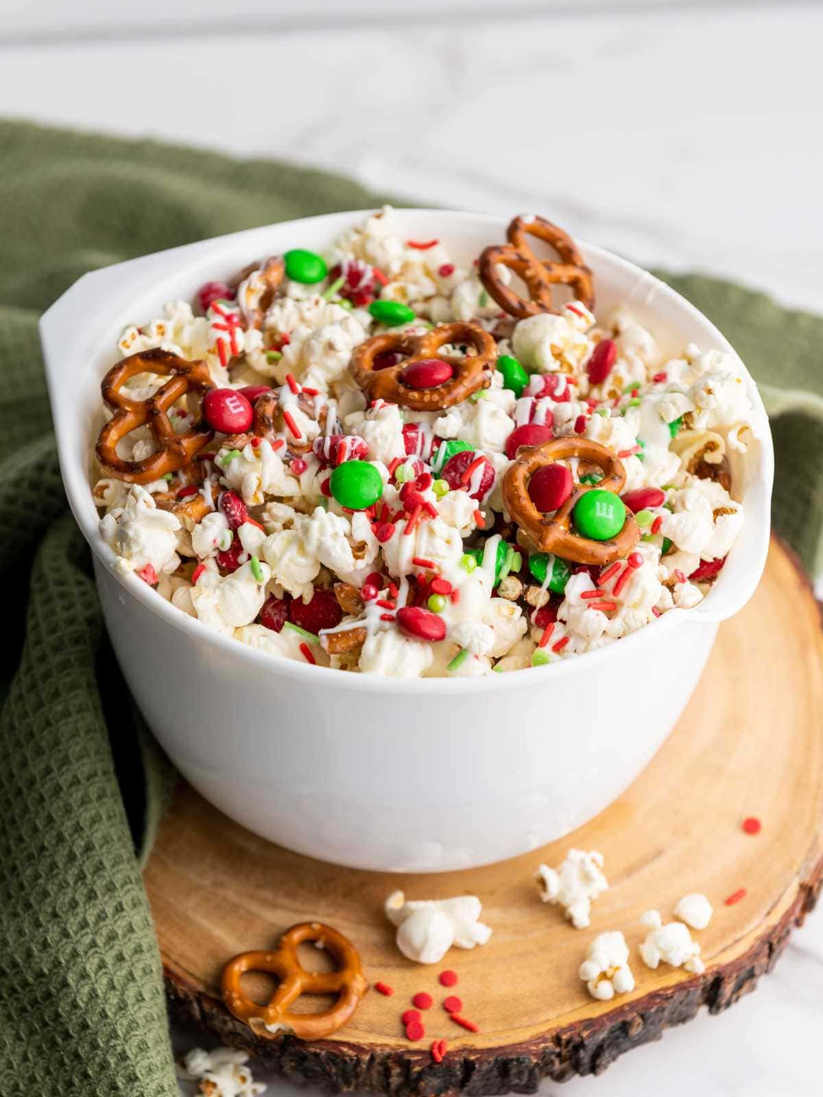 A bowl of popcorn mixed with pretzels, red and green candies, sprinkles, and drizzled with white chocolate.