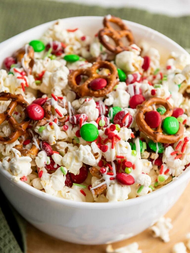 A bowl of popcorn mixed with pretzels, red and green candies, sprinkles, and drizzled with white chocolate.