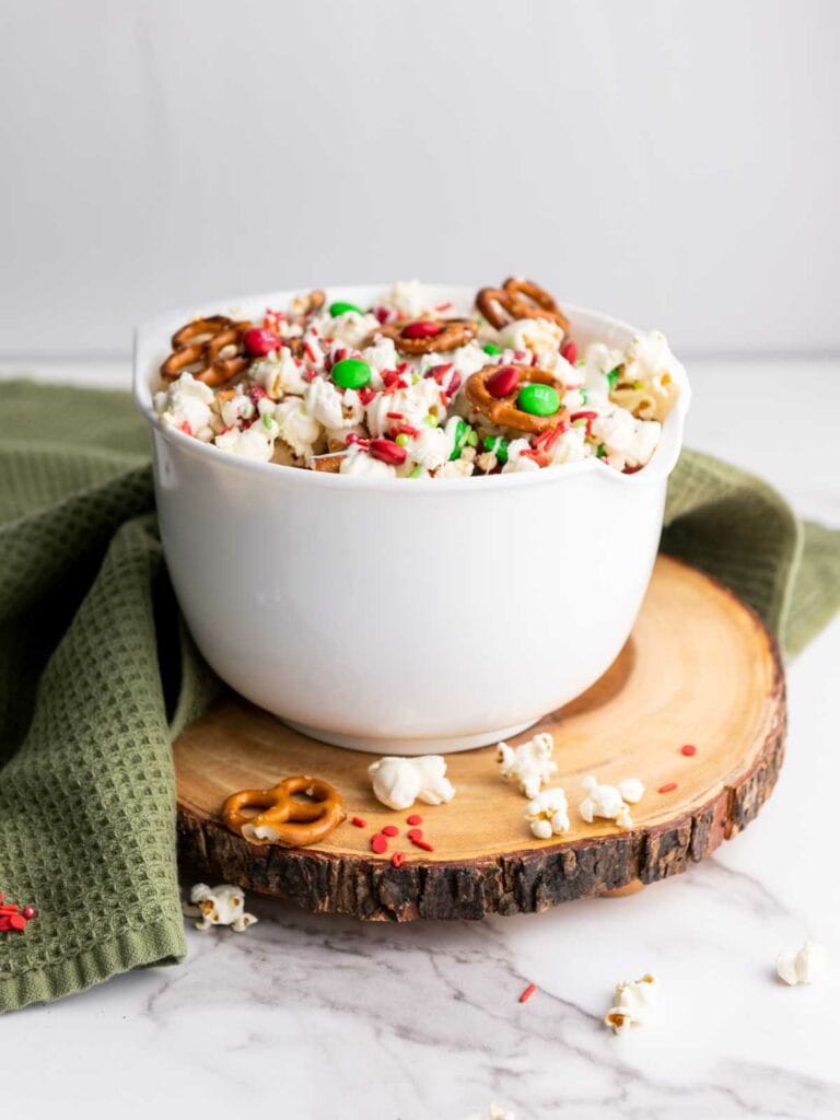 A white bowl filled with popcorn, pretzels, red and green candies, and sprinkles sits on a wooden board with a green cloth underneath.