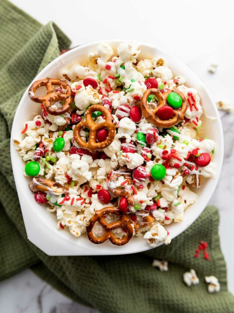 A bowl of popcorn mixed with pretzels, green and red candies, and festive sprinkles on a green textured cloth.