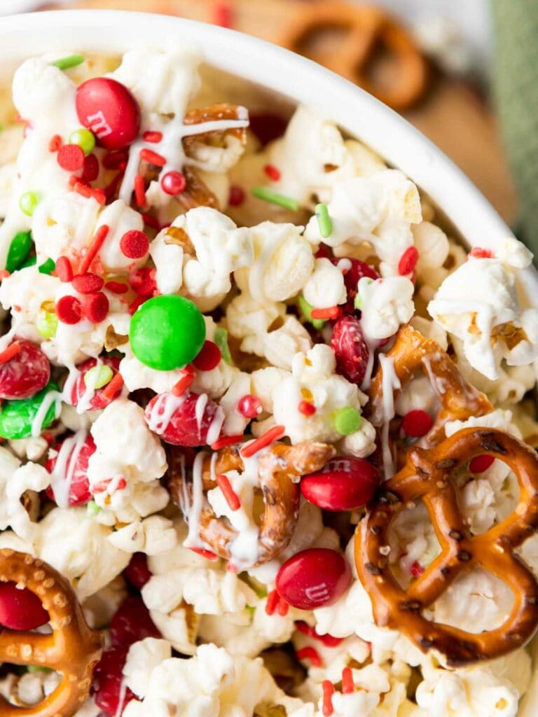 A close-up of a snack mix with popcorn, pretzels, red and green candy, and drizzled white chocolate in a white bowl.