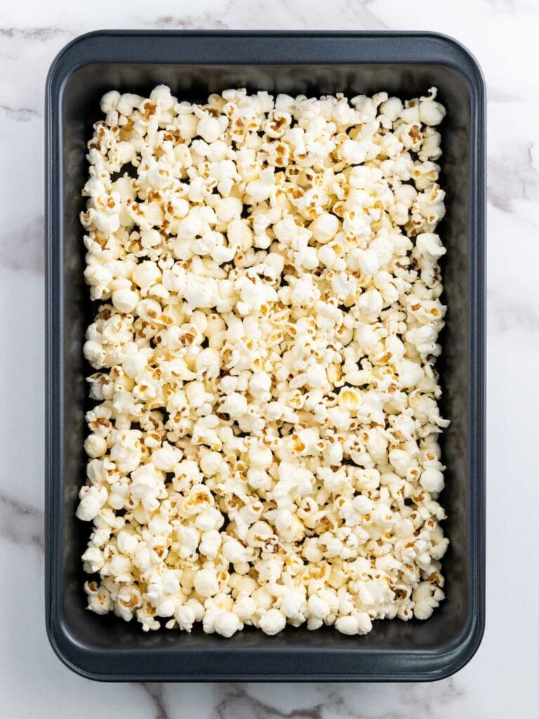 A black rectangular baking tray filled with freshly popped popcorn, placed on a marble surface.