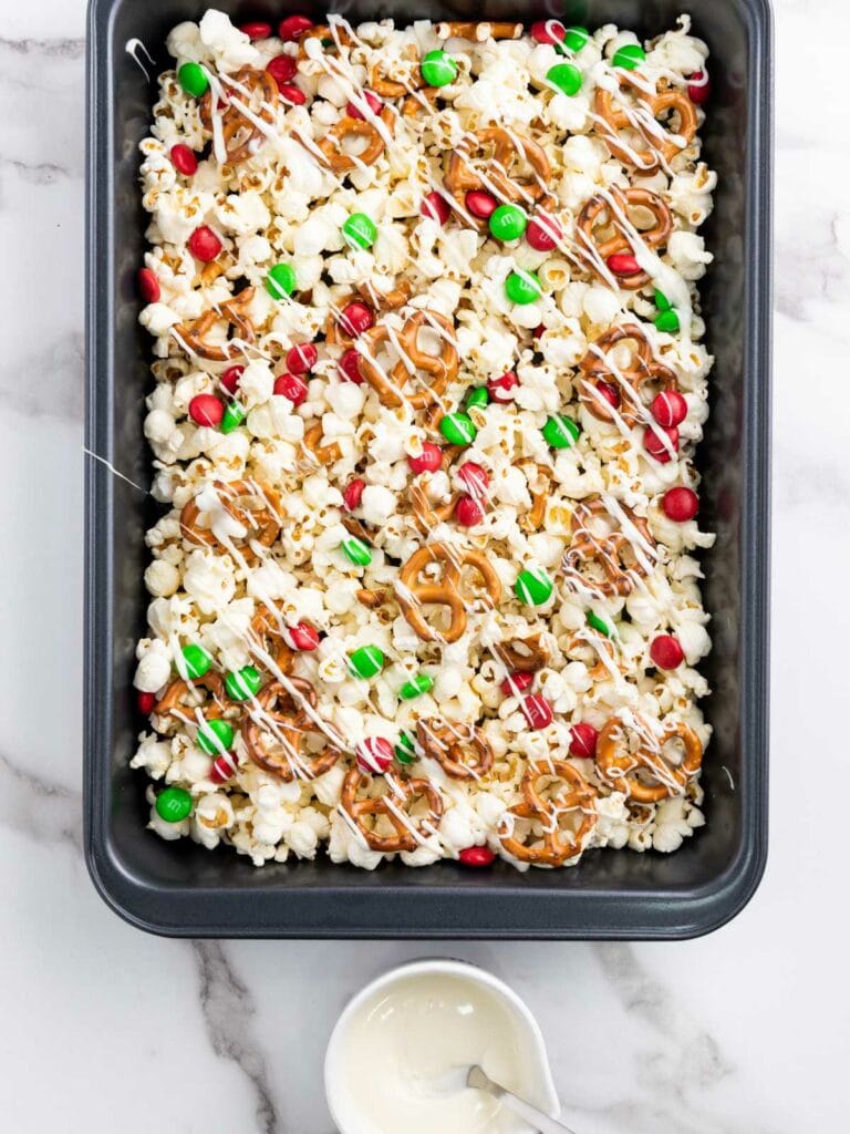 A baking tray of popcorn topped with red and green candies, pretzels, and white chocolate drizzle on a marble surface. A small bowl with melted white chocolate is nearby.