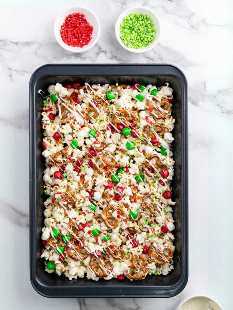 A baking tray with a mix of popcorn, pretzels, candy-coated chocolates, and drizzled white chocolate. Small bowls of red and green sprinkles are on the side.