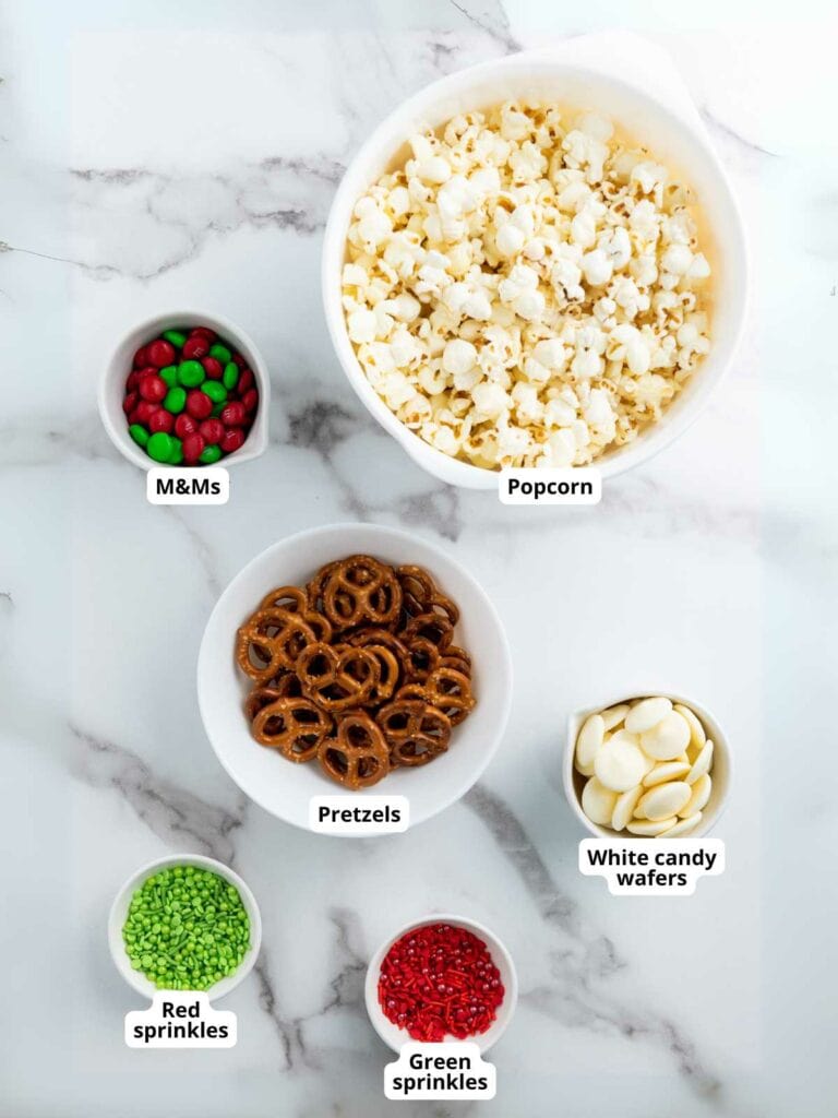 Bowls of popcorn, M&Ms, pretzels, white candy wafers, green sprinkles, and red sprinkles on a marble surface.