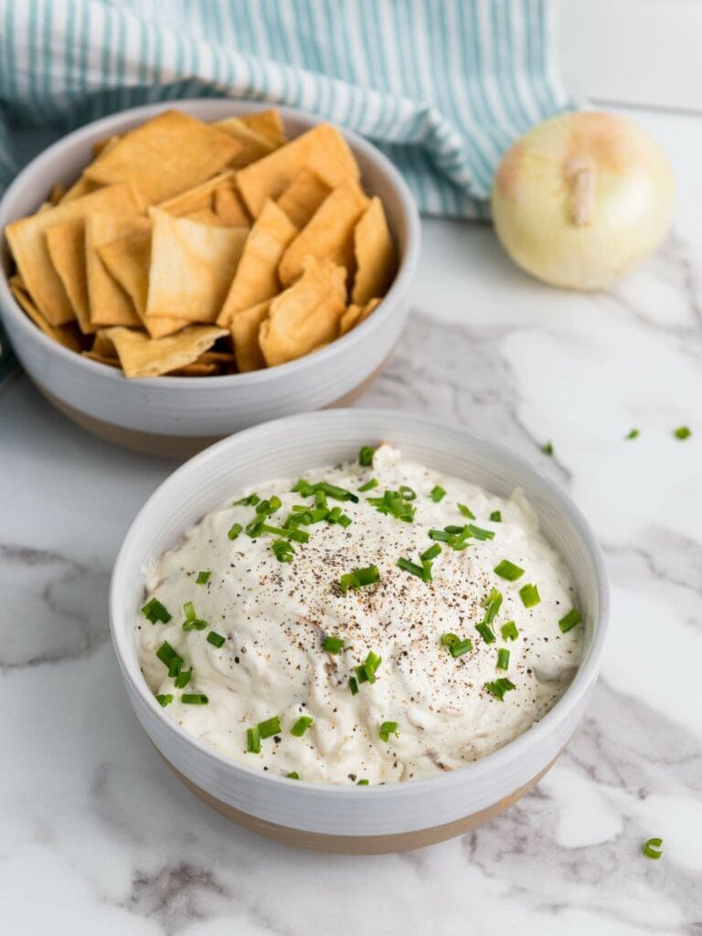 A bowl of creamy french onion dip garnished with chopped green onions and black pepper, next to a bowl of square crackers.