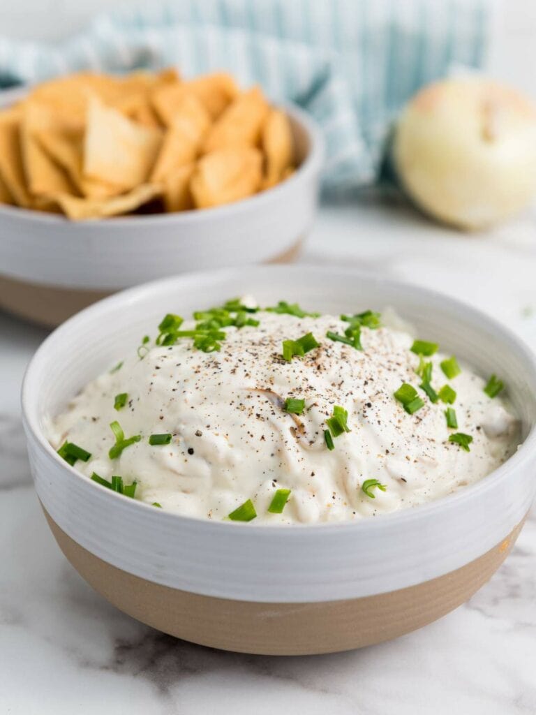 A bowl of creamy french onion dip topped with chopped green onions and black pepper, placed on a marble surface.