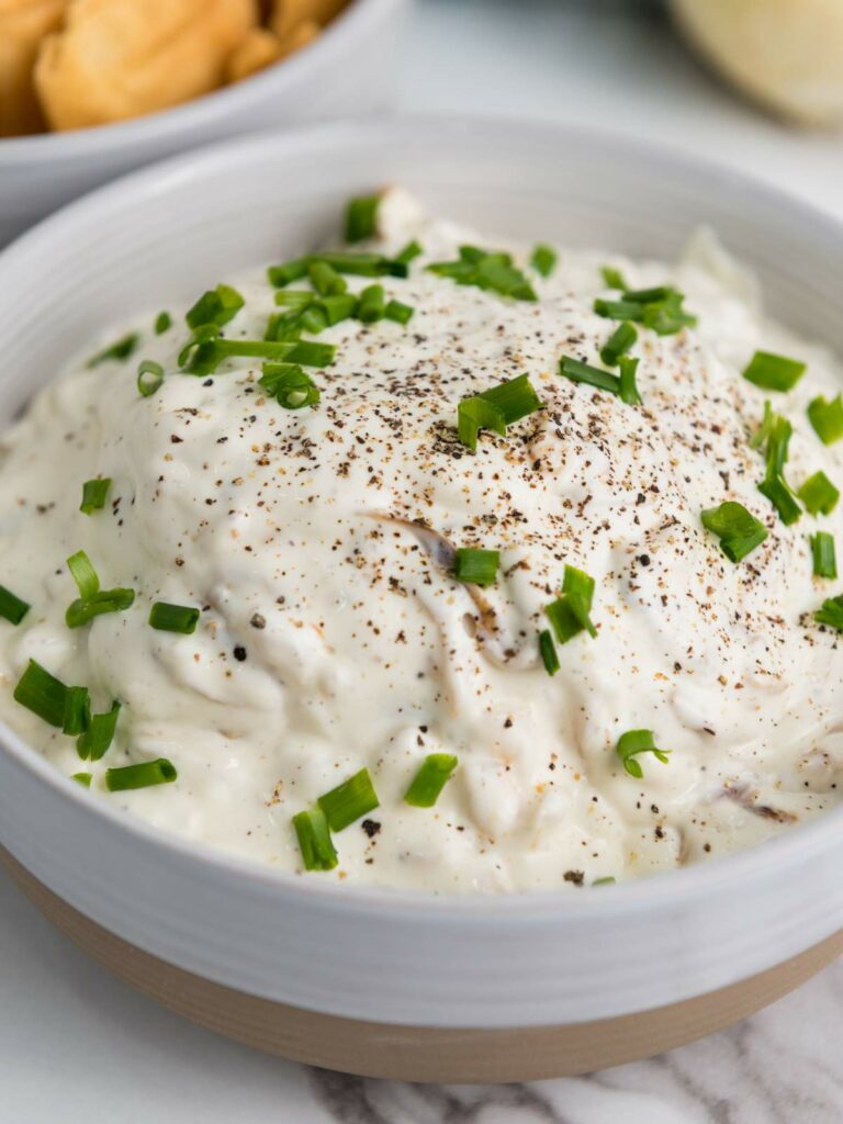 Bowl of creamy french onion dip topped with chopped green onions and pepper, next to a bowl of chips.